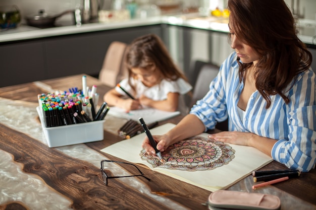 Young beautiful mother and cute daughter draw a mandala pattern together artistic markers and colore...