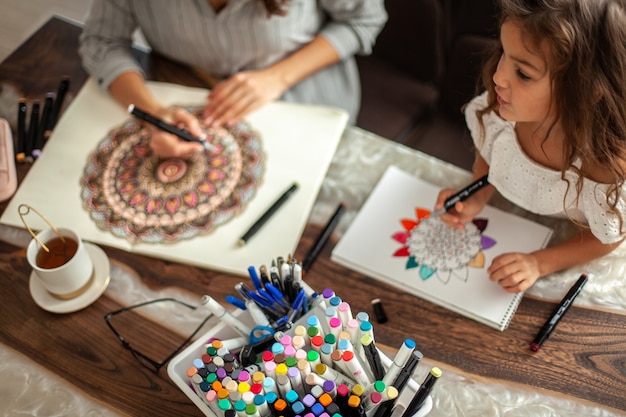 Young beautiful mother and cute daughter draw a mandala pattern together artistic markers and colore...