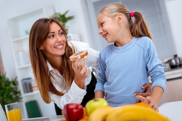 Young beautiful mother and her cute daughter having breakfast in the kitchen. Mother is feeding daughter with slice of bread with pate.