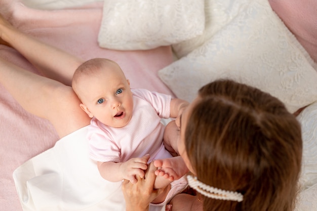 A young beautiful mother holds her daughter a girl of 6 months on her lap on a white bed, playing and kissing her