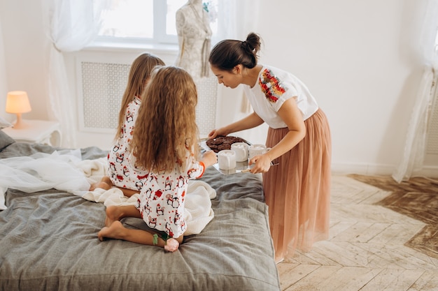 Young beautiful mother is bringing cocoa with Marshmallows and cookies to her daughters sitting on the bed in the full of light cozy room .