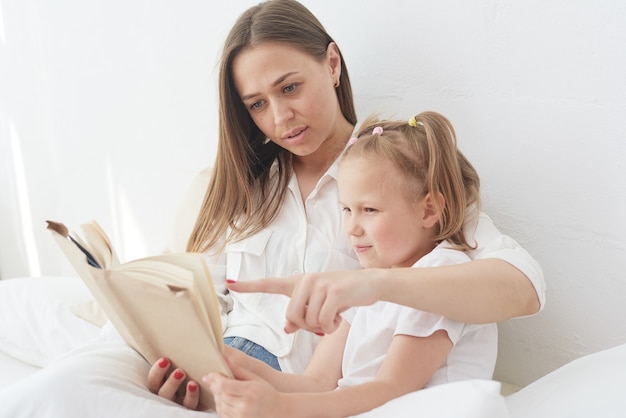 Young beautiful mother is reading a book to her daughter. Happy family, mother and daughter reading a book while lying in bed, smiling nanny mom telling funny story to cute preschool girl.