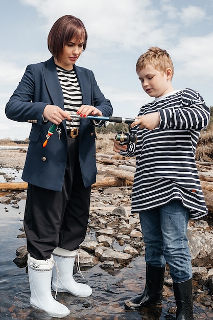 Young, beautiful mother and son have fun fishing on the lake. Mom and son in striped vests on the river bank with a fishing rod.