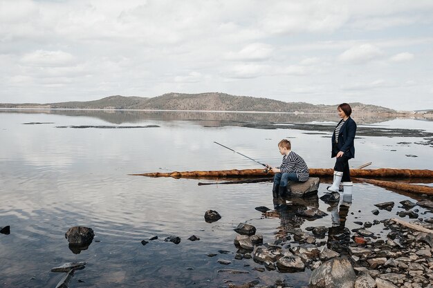 Young, beautiful mother and son have fun fishing on the lake. Mom and son in striped vests on the river bank with a fishing rod.