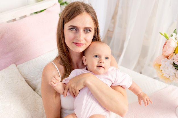 A young beautiful mother with a 6-month-old daughter in her arms sits on a white bed in flowers and hugs her, a place for text