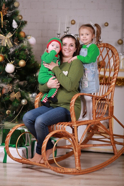 Young beautiful mother with her baby son in elf costume and little girl sitting in rocking chair near christmas tree.