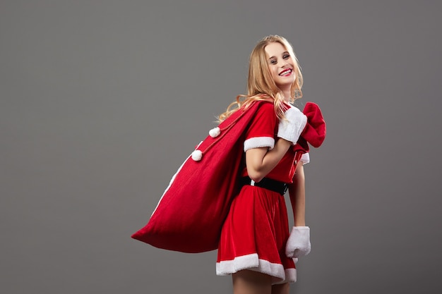 Young and beautiful mrs.Santa Claus dressed in the red robe and white gloves holds the bag with Christmas gifts on her back. .