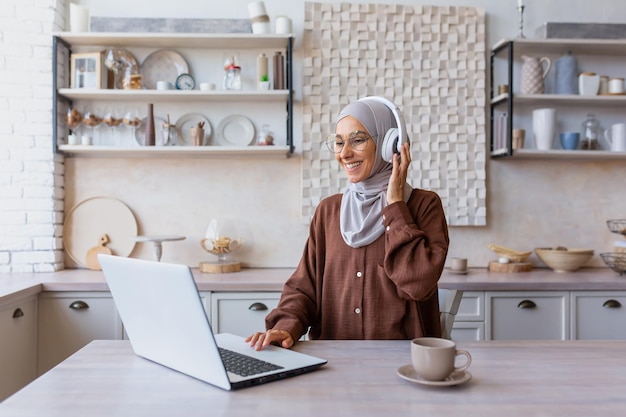 Young beautiful muslim student woman in hijab sitting at home in kitchen wearing headphones and