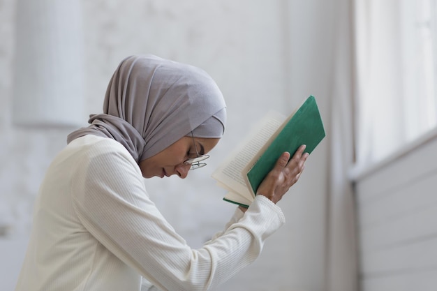 A young beautiful muslim woman in a hijab is praying at home sitting by the window