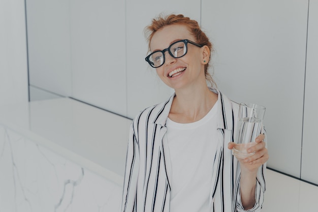 Young beautiful pleased redhaired woman having healthy habits drinking mineral water in morning