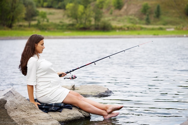 Young beautiful pregnant woman fishing on the river bank