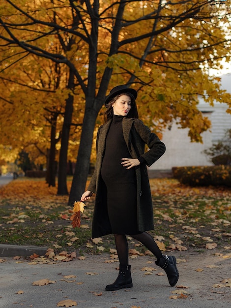 Young beautiful pregnant woman in a hat with dark hair in a black tight dress and coat posing