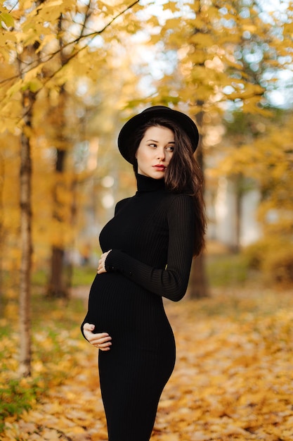 Young beautiful pregnant woman in a hat with dark hair in a black tight dress posing