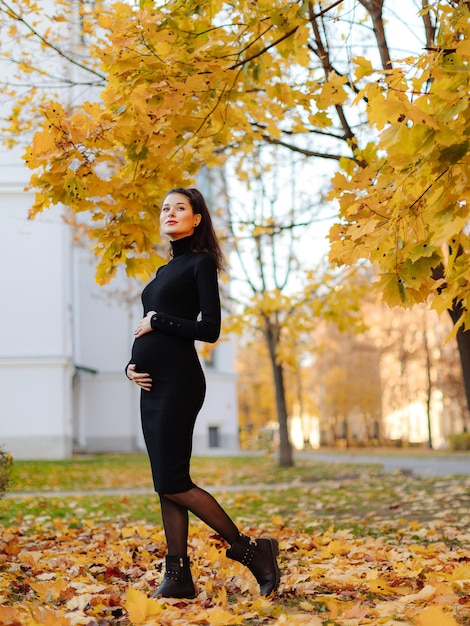 Young beautiful pregnant woman with dark hair in a black tight dress posing on an autumn