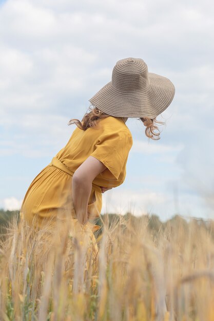 A young beautiful pregnant woman in a yellow dress and hat walks through a wheat orange field on a sunny summer day