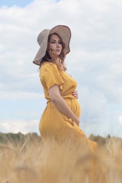 A young beautiful pregnant woman in a yellow dress and hat walks through a wheat orange field on a sunny summer day