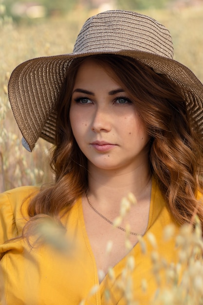 A young beautiful pregnant woman in a yellow dress and hat walks through a wheat orange field on a sunny summer day