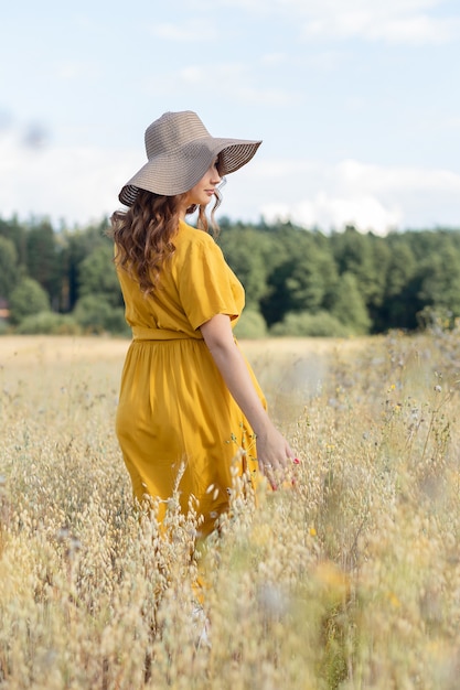 A young beautiful pregnant woman in a yellow dress and hat walks through a wheat orange field on a sunny summer day