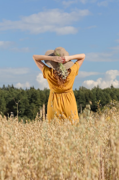 A young beautiful pregnant woman in a yellow dress and hat walks through a wheat orange field on a sunny summer day