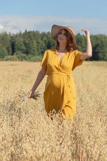 A young beautiful pregnant woman in a yellow dress and hat walks through a wheat orange field on a sunny summer day