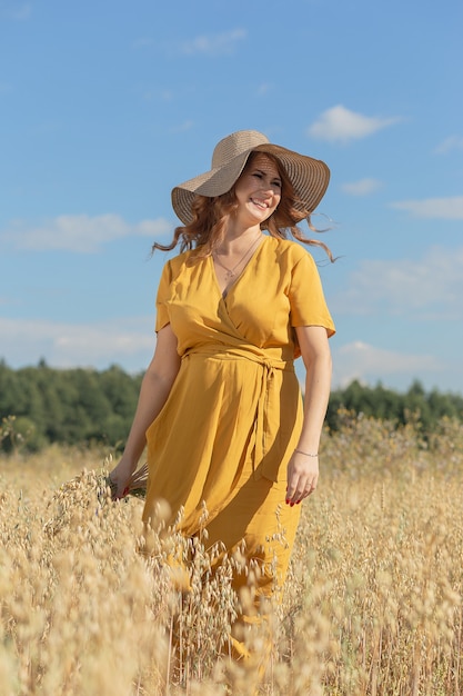 A young beautiful pregnant woman in a yellow dress and hat walks through a wheat orange field on a sunny summer day