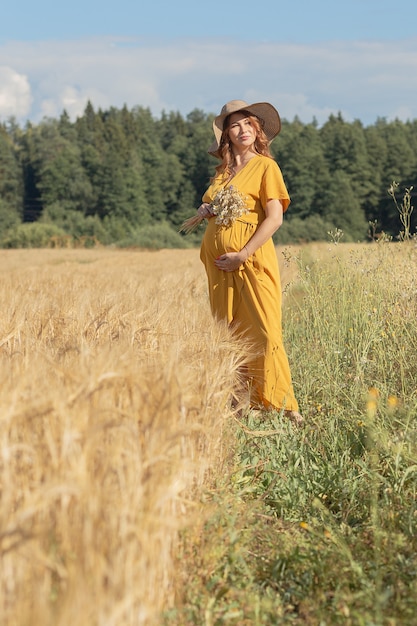 A young beautiful pregnant woman in a yellow dress and hat walks through a wheat orange field on a sunny summer day