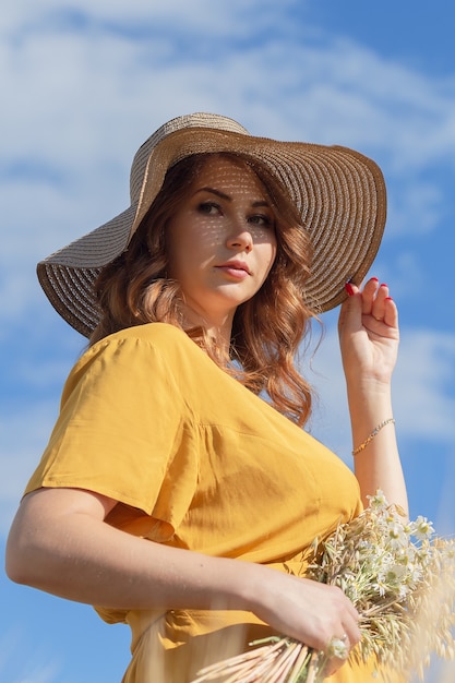 A young beautiful pregnant woman in a yellow dress and hat walks through a wheat orange field on a sunny summer day