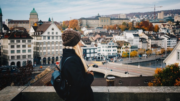 Young beautiful pretty blonde tourist girl in warm hat and coat with backpack walking at cold autumn in Europe city enjoying her travel in Zurich Switzerland