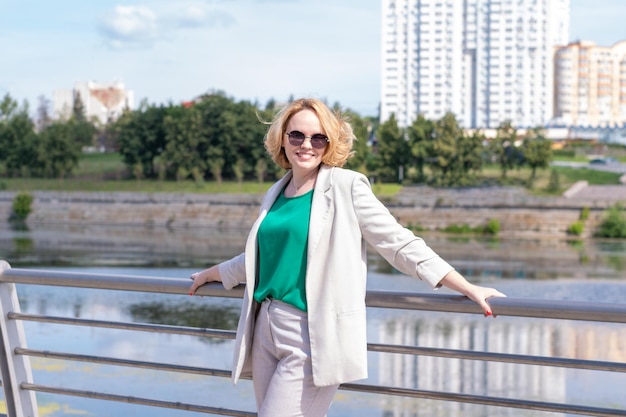 A young beautiful pretty girl in a suit jacket is standing on the bank of the river holding on to the railing