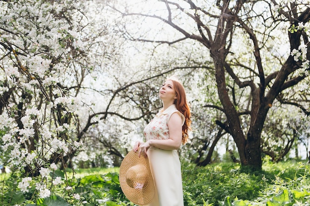 Young beautiful red-haired girl walks in a spring blooming apple orchard