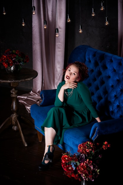 Young beautiful red-haired woman with professional makeup in green dress posing in luxurious interior on blue sofa