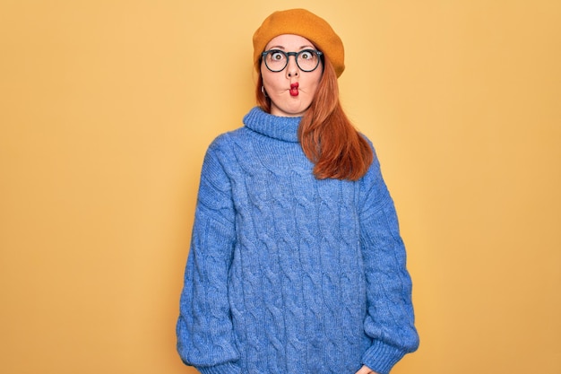 Young beautiful redhead woman wearing french beret and glasses over yellow background making fish face with lips crazy and comical gesture Funny expression