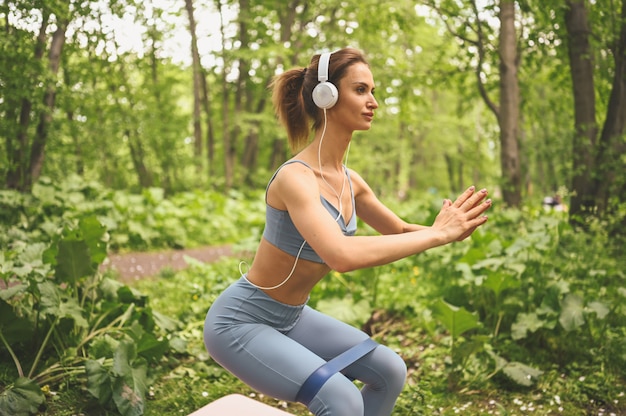 Young beautiful slim fit girl in blue sportswear with big white headphones doing exercise with fitness training gum in the park