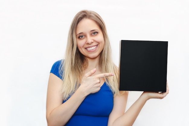 A young beautiful smiling blonde woman in a blue tshirt holds a square black box and points to it on a white wall