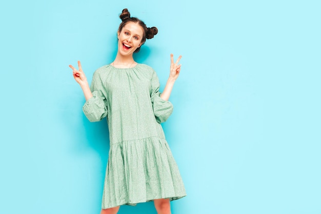 Young beautiful smiling female in trendy summer green dress Sexy carefree woman with two horns hairstyle posing near blue wall in studio Positive model having fun Shows peace sign