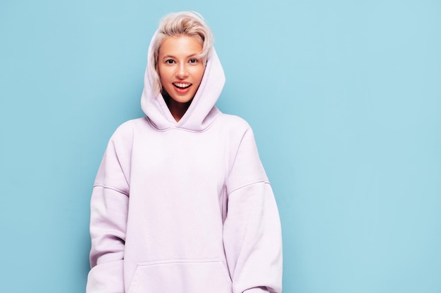 Young beautiful smiling female in trendy summer hoodie Sexy carefree woman posing near blue wall in studio Positive blond model having fun and going crazy Cheerful and happy