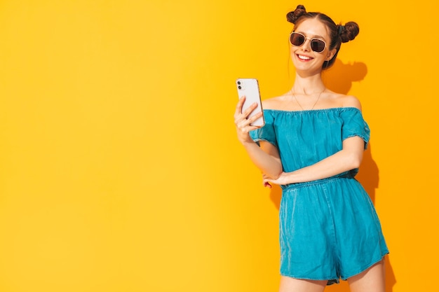 Young beautiful smiling female in trendy summer jeans overalls Sexy carefree woman with two horns hairstyle posing near yellow wall in studio Positive model having fun Isolated Taking selfie