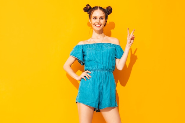 Young beautiful smiling female in trendy summer jeans overalls Sexy carefree woman with two horns hairstyle posing near yellow wall in studio Positive model having fun Isolated