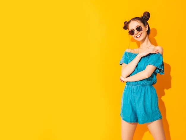 Young beautiful smiling female in trendy summer jeans overalls Sexy carefree woman with two horns hairstyle posing near yellow wall in studio Positive model having fun Isolated