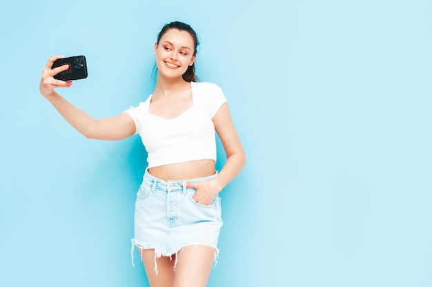 Young beautiful smiling female in trendy summer jeans skirt and top clothes Sexy carefree woman posing near blue wall in studio Positive brunette model having fun Taking selfie