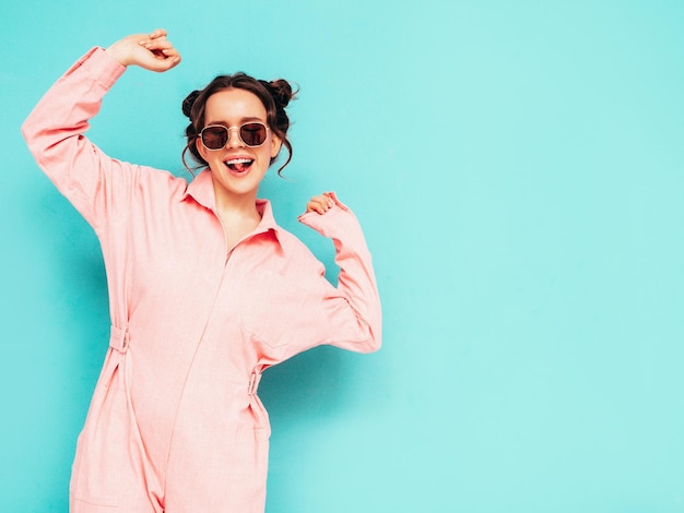 Photo young beautiful smiling female in trendy summer pink overalls sexy carefree woman posing near blue wall in studio positive model having fun and going crazy cheerful and happy dancing in sunglasses