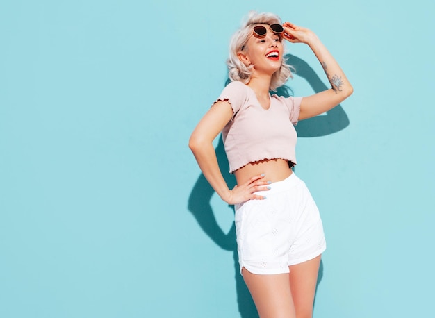 Young beautiful smiling female in trendy summer shorts and top clothes Sexy carefree woman posing near blue wall in studio Positive blond model having fun and going crazy Cheerful and happy