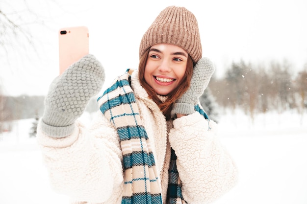 Young beautiful smiling hipster female in trendy warm clothes and scarf.Carefree woman posing in the street in park. Positive pure model having fun in snow. Enjoying winter moments.Taking selfie