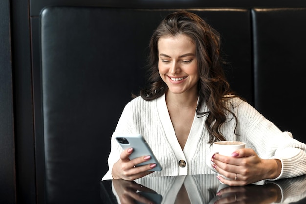 Young beautiful smiling woman holding coffee cup and looking at smartphone