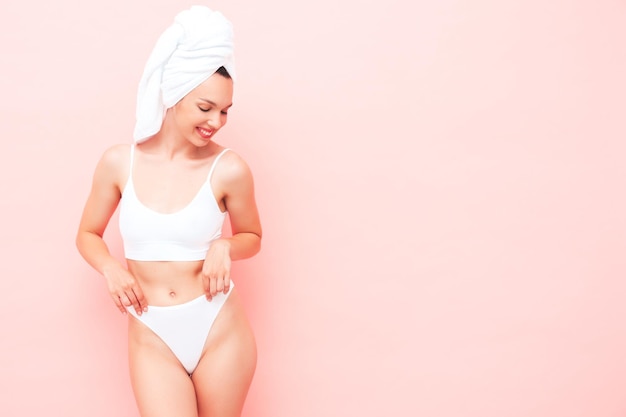 Young beautiful smiling woman in white lingerie. Sexy carefree model in underwear and towel on head posing pink wall in studio. Positive and happy female enjoying morning