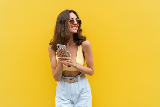 Young beautiful smiling woman with mobile phone Carefree woman posing in the street near yellow wall Positive model outdoors in sunglasses Happy and excited