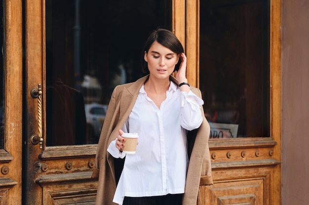 Young beautiful stylish businesswoman with coffee to go dreamily standing outdoor