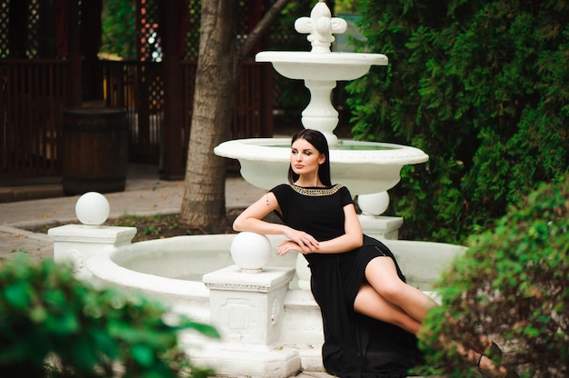 Young beautiful stylish girl walking and posing in short black dress in city near fountains. Outdoor summer portrait of young classy woman.