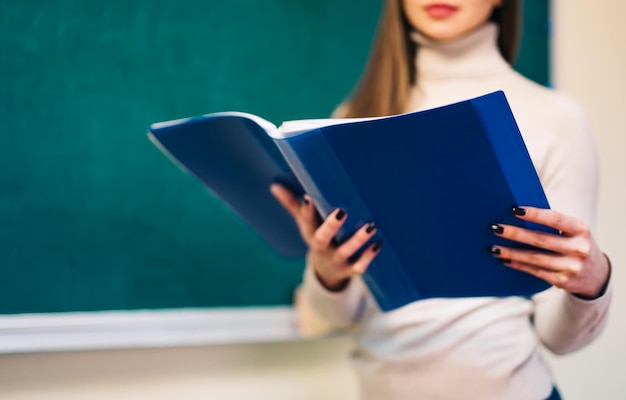 Young beautiful teacher standing near the board and looking at the opening book in classroom Cropped view of the tutor in classroom of school or university Education and job concept