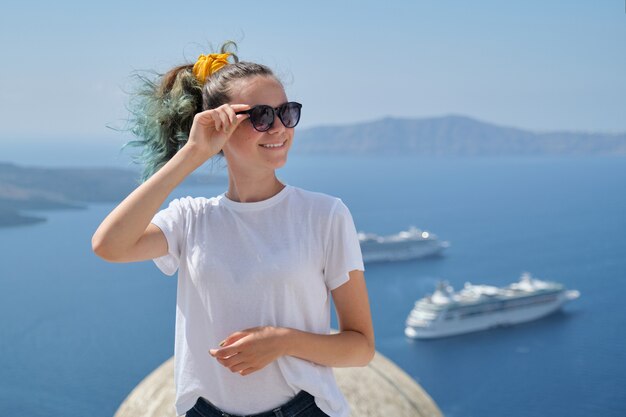 Young beautiful teenager girl tourist posing smiling, background summer sunny scenic sea landscape with white cruise liners in Aegean Sea on famous Greek island Santorini, copy space